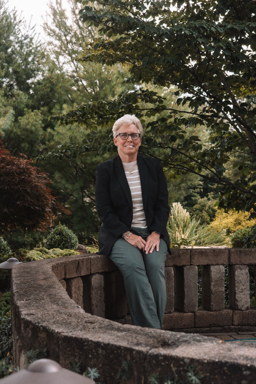 Debra Shumar seated on a stone garden wall, smiling, surrounded by mature garden plantings.